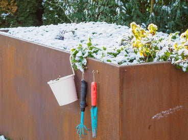 A covered raised beds winter signified by the layer of snow on the bed, with some tools dangling from the side of the raised bed