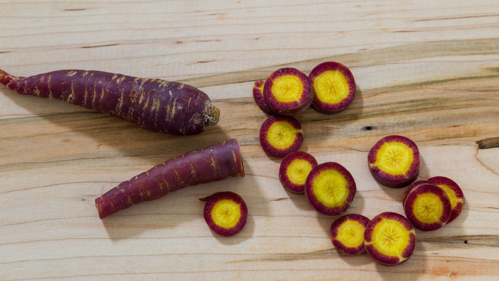 An overhead and close-up shot of sliced Cosmic Purple Carrots, showcasing its purple skin and golden-yellow interiors, all placed on a wooden surface indoors